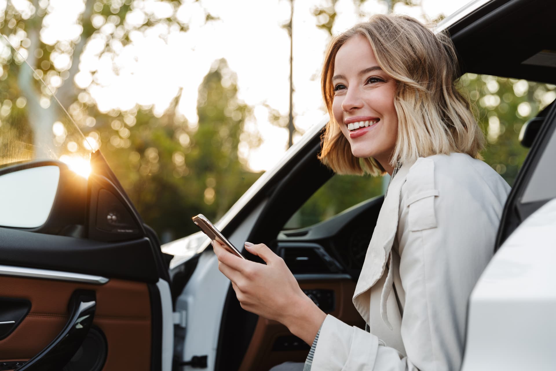 A driver smiling with a phone beside her car